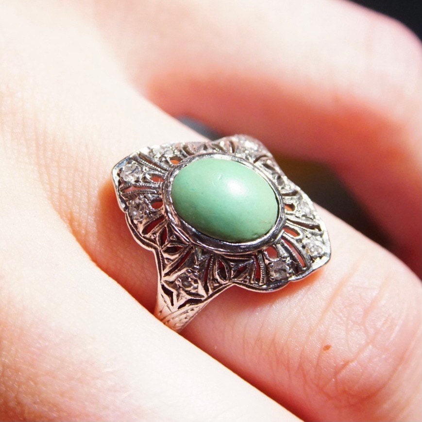 Vintage 18K white gold art deco filigree ring with a matte green turquoise stone, surrounded by intricate diamond encrusted shield design, shown on a finger, size 7 3/4 US.