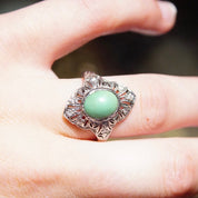 Vintage 18K white gold turquoise ring with Art Deco filigree design and diamond encrusted shield-shaped setting holding a matte green turquoise stone, shown on a hand, ring size 7 3/4 US.