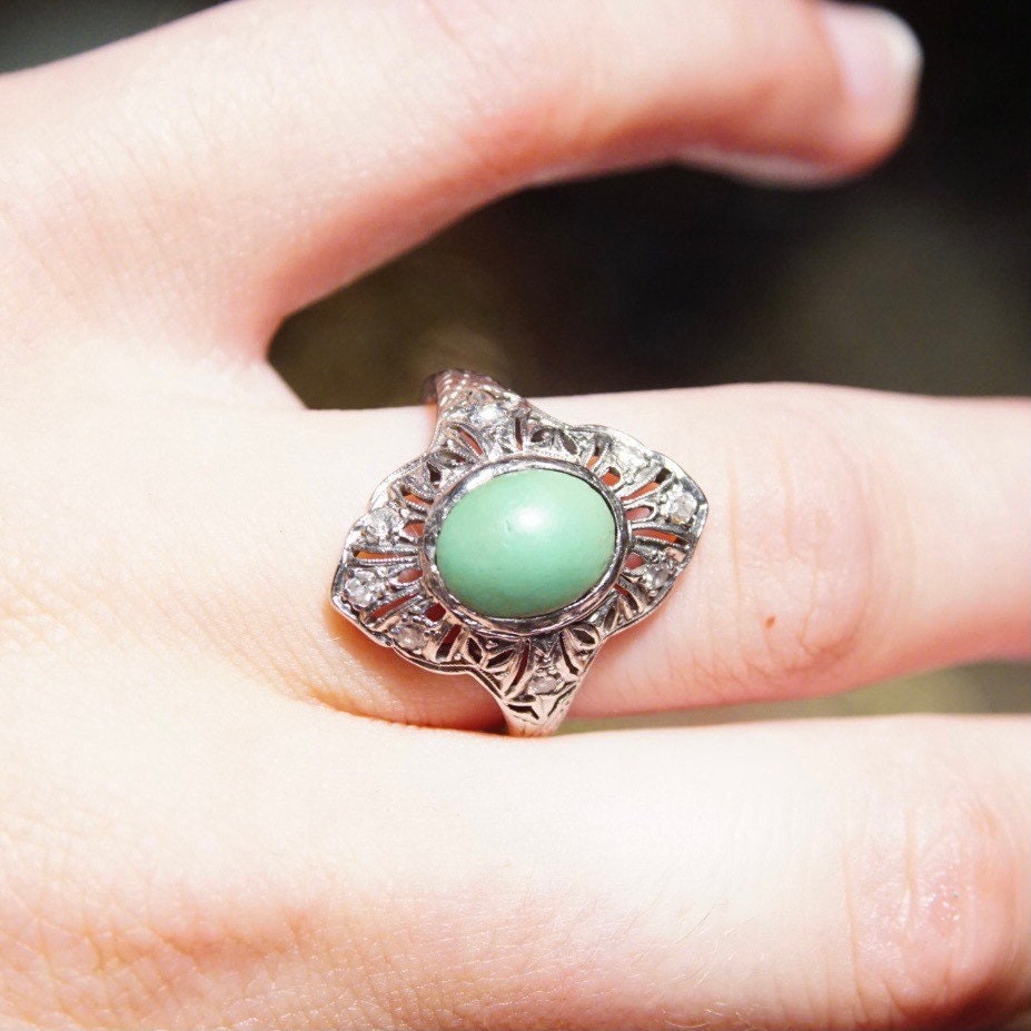 Vintage 18K white gold turquoise ring with Art Deco filigree design and diamond encrusted shield-shaped setting holding a matte green turquoise stone, shown on a hand, ring size 7 3/4 US.