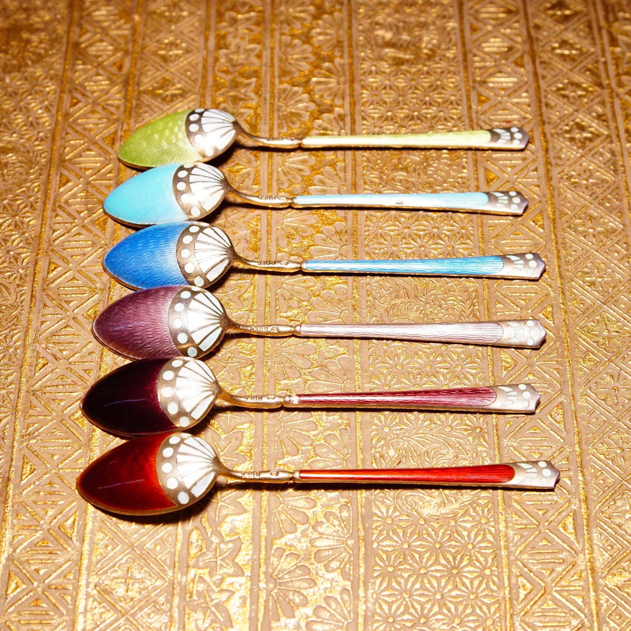 Antique Art Deco sterling silver guilloche enamel demitasse spoon set of 6, featuring colorful decorative tea spoons in shades of blue, green, purple and red, Scandinavian origin, marked 930S, displayed on an ornate gold patterned background.