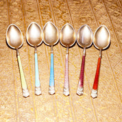Alt text: Six antique sterling silver demitasse spoons with colorful guilloche enamel handles in turquoise, white, and red, laid out in a row on an ornate gold patterned surface.