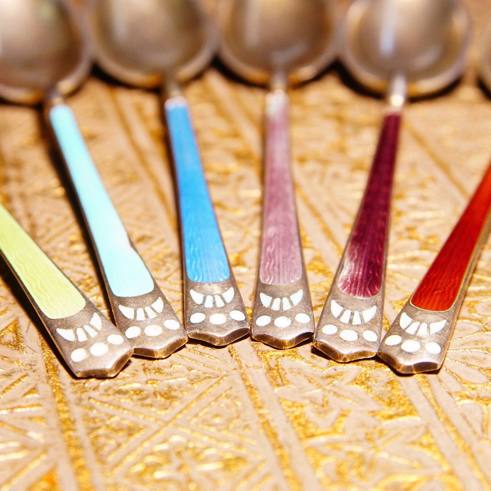 Antique Art Deco sterling silver guilloche enamel demitasse spoon set with colorful handles in blue, turquoise, red, and purple, arranged in a decorative fan pattern on a textured gold background.