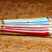 Alt text: Close-up view of six antique sterling silver demitasse spoons with colorful guilloche enamel handles in red, light blue, and dark blue, resting on an ornate gold patterned surface.