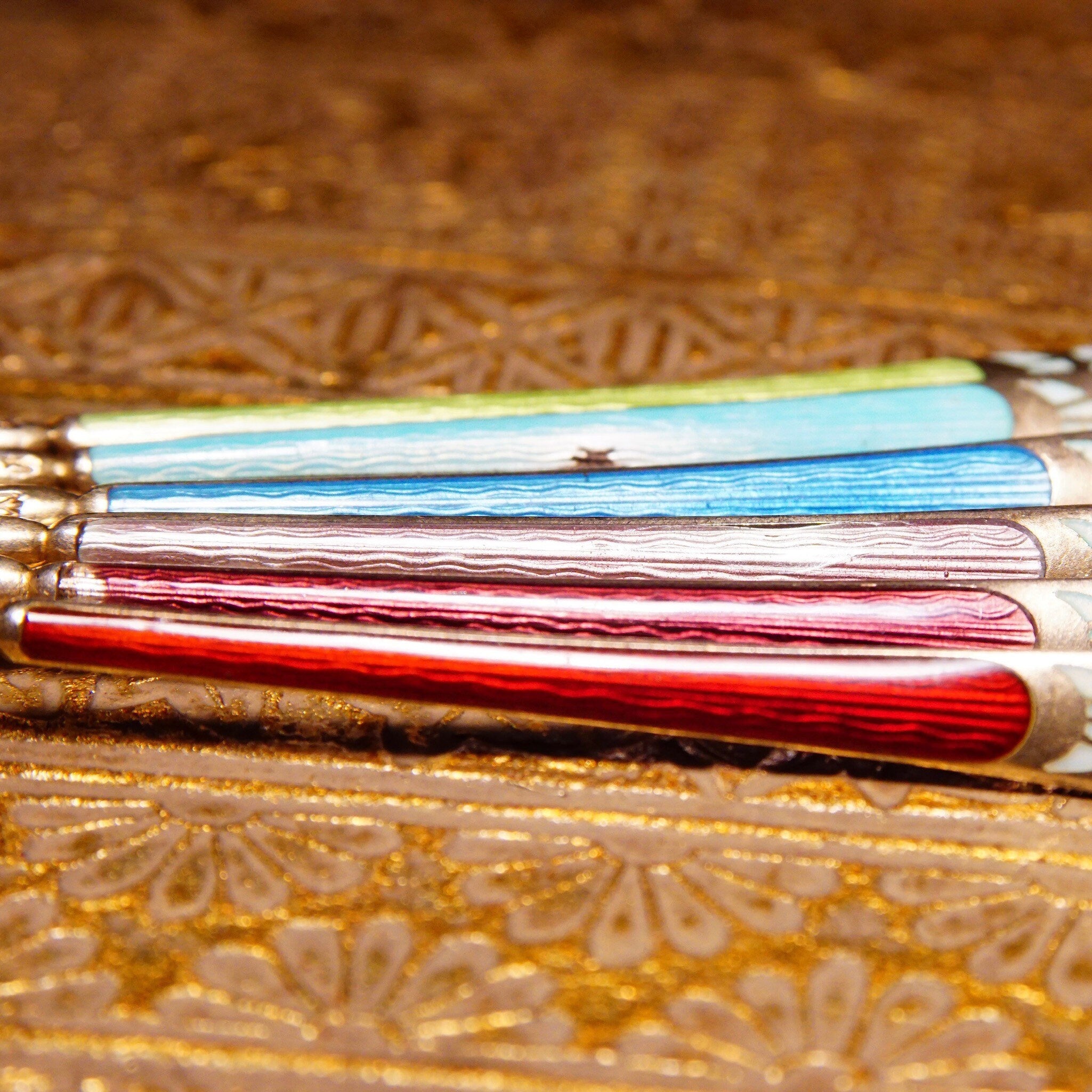 Alt text: Close-up view of six antique sterling silver demitasse spoons with colorful guilloche enamel handles in red, light blue, and dark blue, resting on an ornate gold patterned surface.