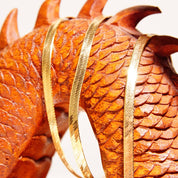 Overlapping shiny copper-colored fish scales or feathers, forming an intricate herringbone pattern that curves across the image, suggesting it may be a closeup of a piece of jewelry or metalwork.