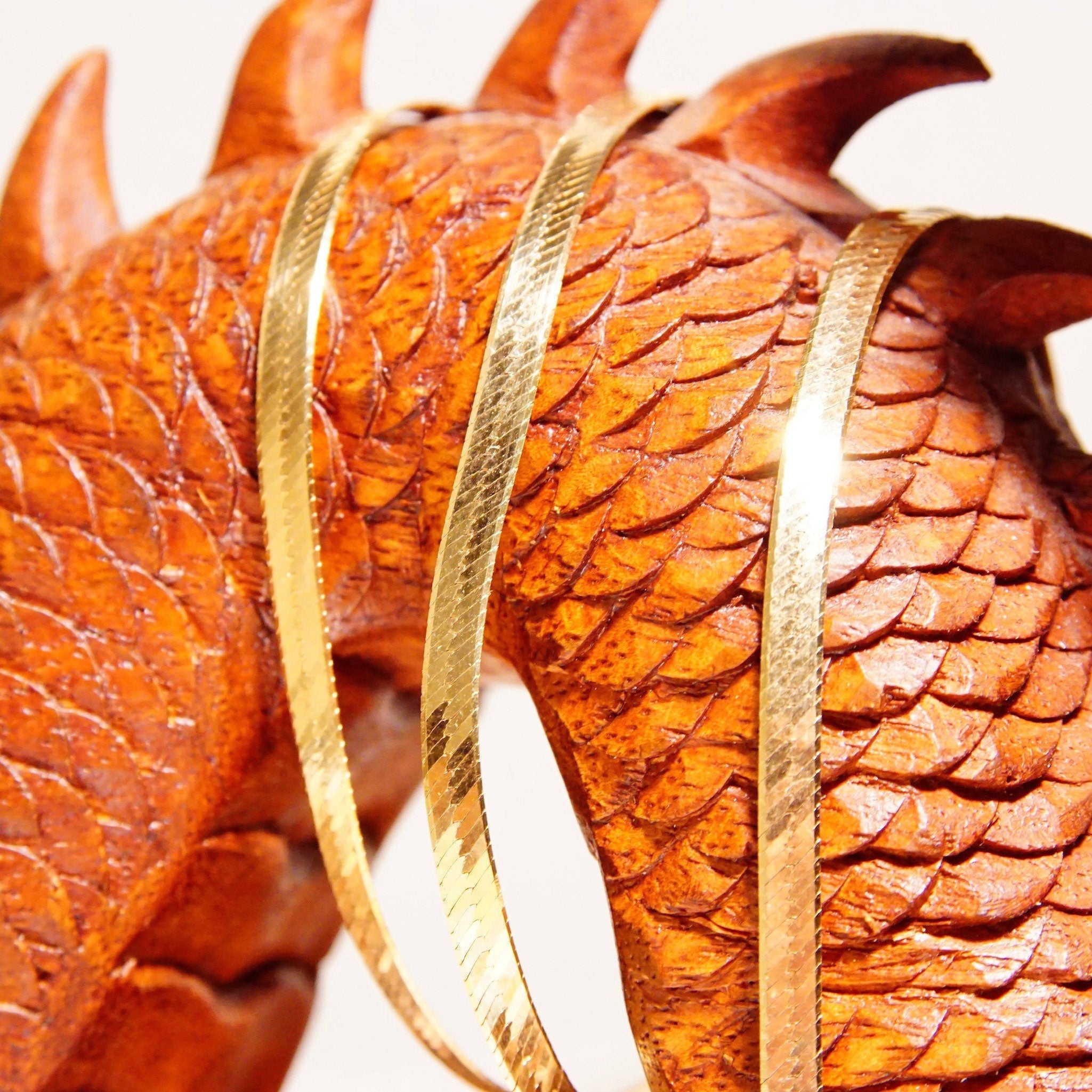 Overlapping shiny copper-colored fish scales or feathers, forming an intricate herringbone pattern that curves across the image, suggesting it may be a closeup of a piece of jewelry or metalwork.