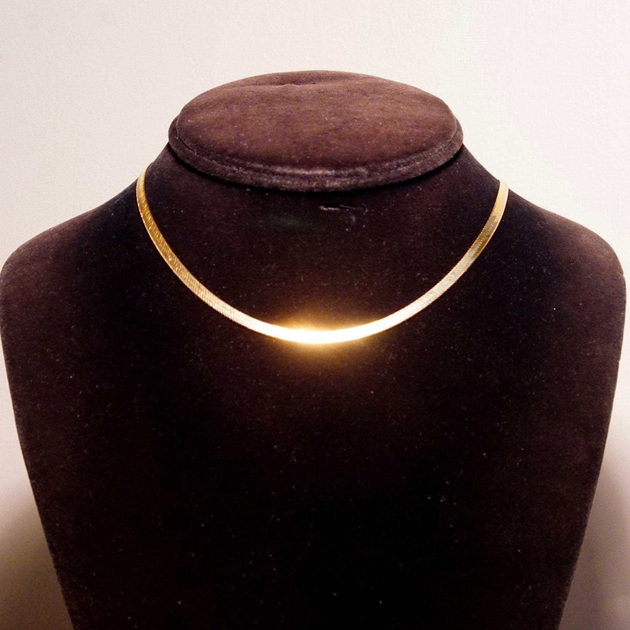 Vintage 14K yellow gold Italian herringbone chain necklace on black velvet bust display, showing the minimalist flat chain design and warm golden glow against the dark background.