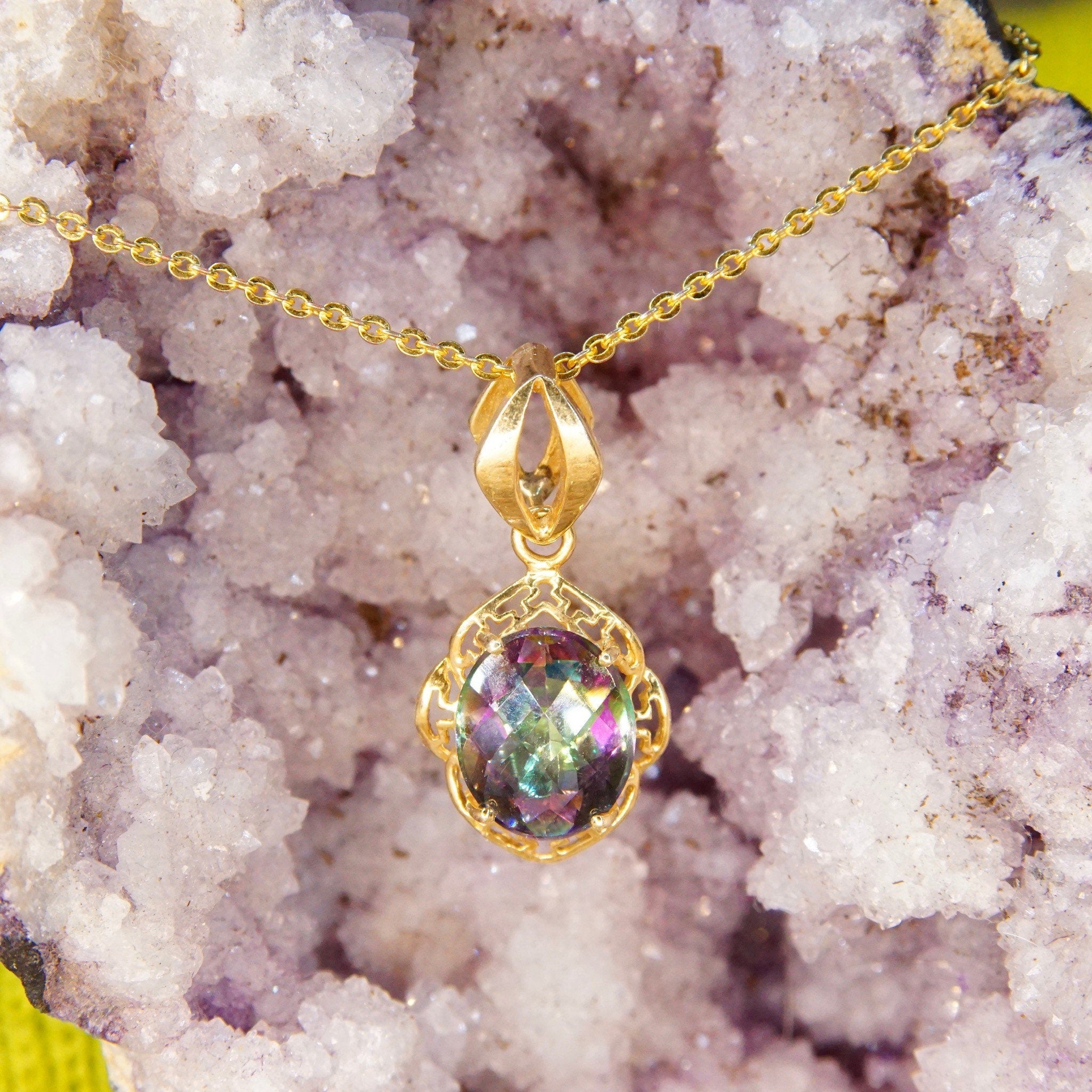 Vintage 14K yellow gold pendant featuring a faceted mystic fire topaz gemstone with purple and green hues, set in an ornate gold setting with a stylized bail, measuring 26mm by 13mm, photographed on colorful crystal background.
