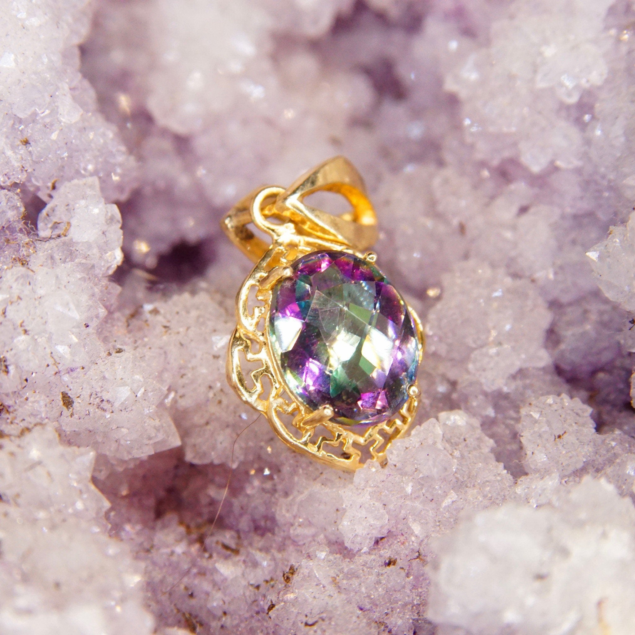 Vintage 14K yellow gold pendant featuring a faceted oval mystic fire topaz gemstone with purple and green hues, set in an ornate gold setting with a stylized bail, measuring approximately 26mm by 13mm, displayed on a background of purple crystal clusters.