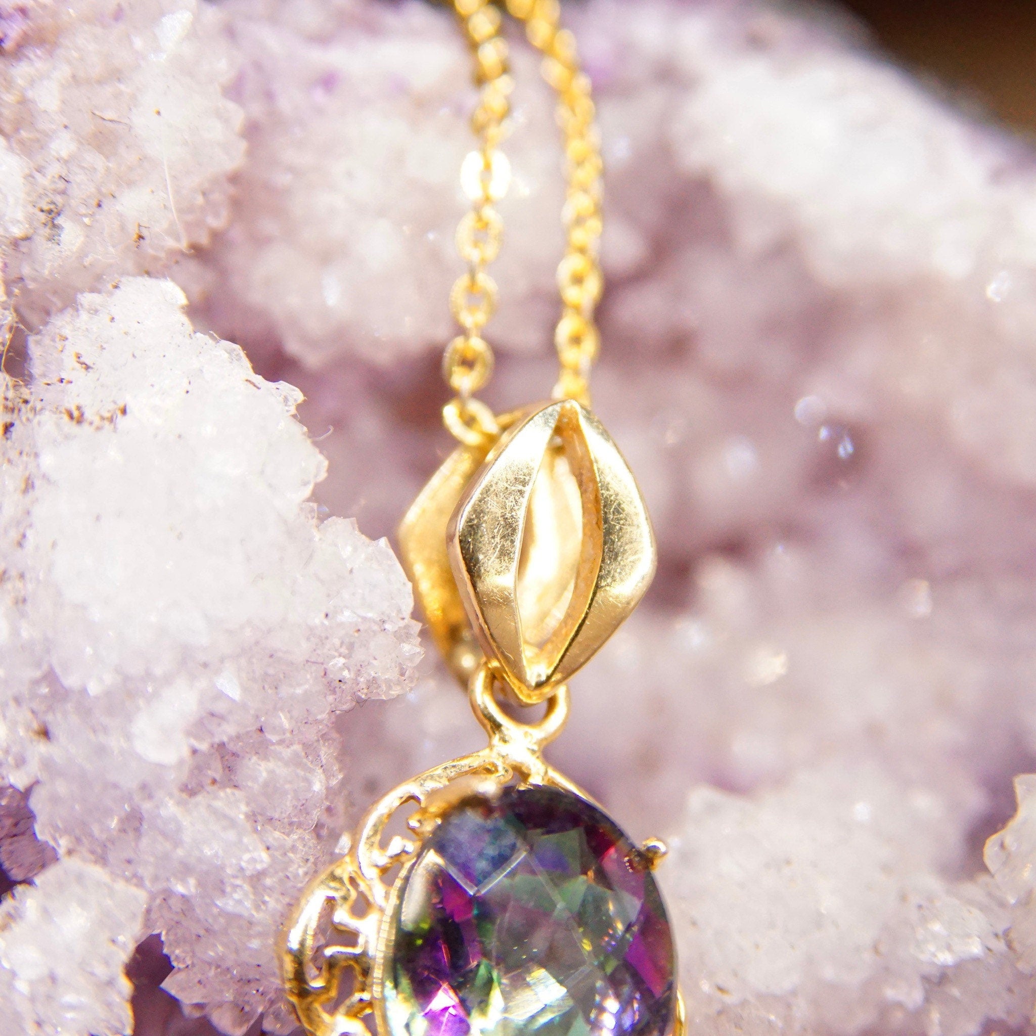 Alt text: Vintage 14K yellow gold pendant necklace featuring an oval faceted mystic fire topaz gemstone with purple and green hues, set in an ornate gold setting with a stylized bail, measuring approximately 26mm by 13mm, photographed on a sparkly pink crystal background.