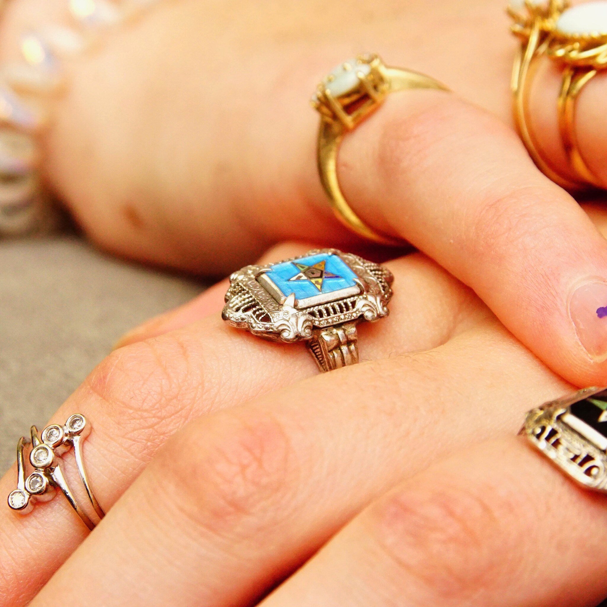 Vintage sterling silver filigree Order of the Eastern Star ring with blue and black enamel detail on fingers, alongside other ornate gold rings.