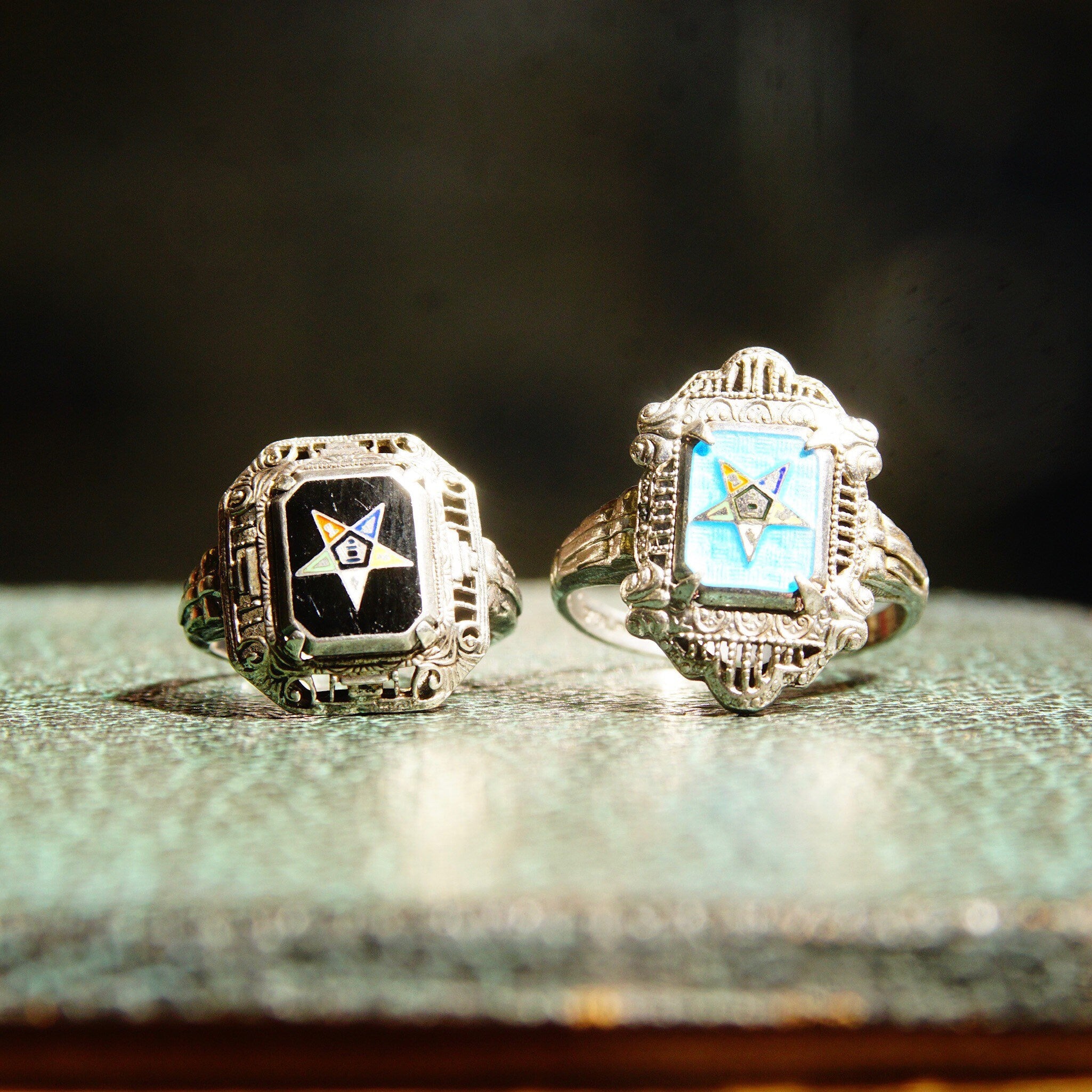Vintage sterling silver filigree Order of the Eastern Star rings with blue and black enamel, featuring Masonic symbol and star designs, displayed on weathered blue fabric.