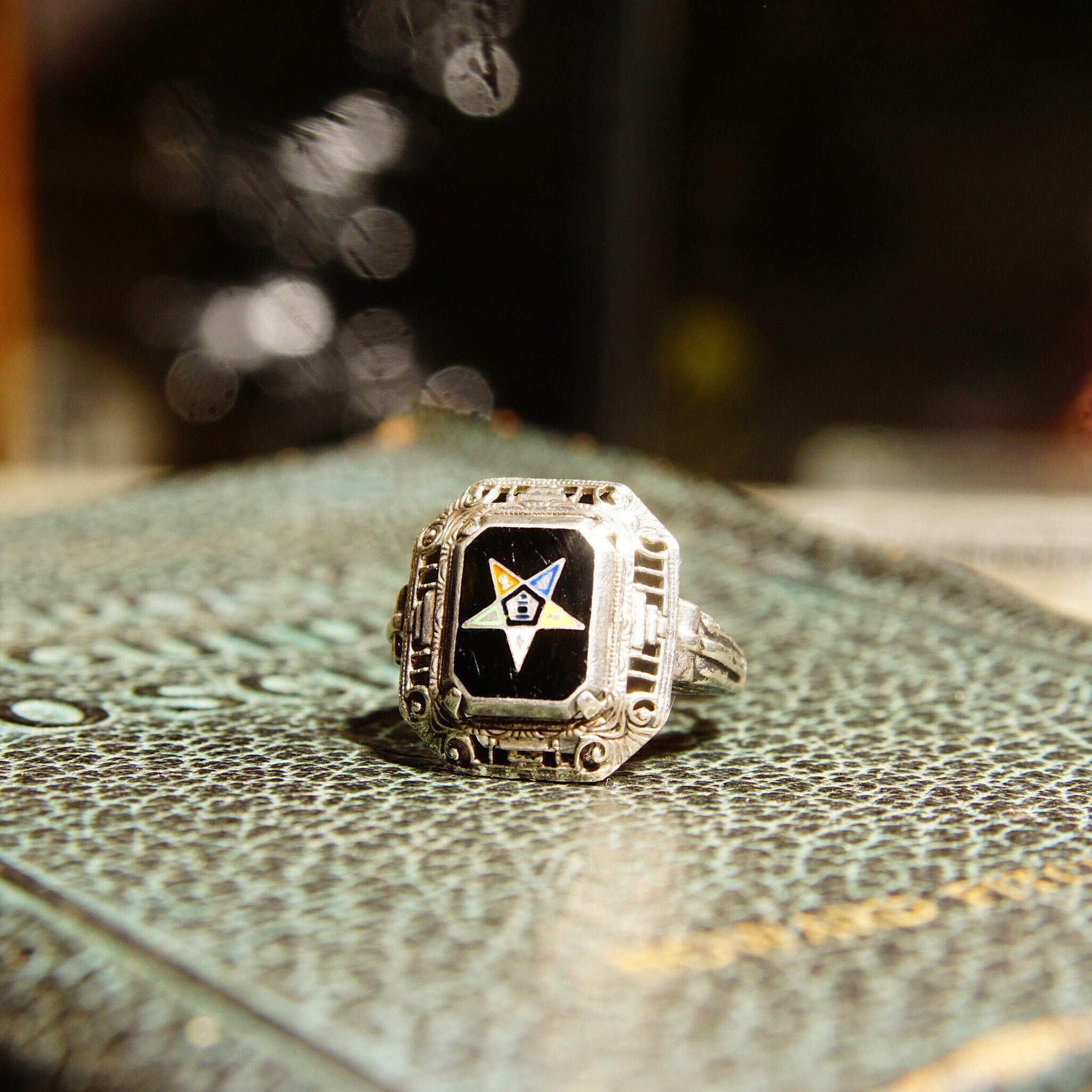 Vintage Sterling Silver Filigree Enamel Order Of The Eastern Star Ring with Black Enamel and Gold Star sitting on an ornate patterned surface with blurred bokeh lights in the background
