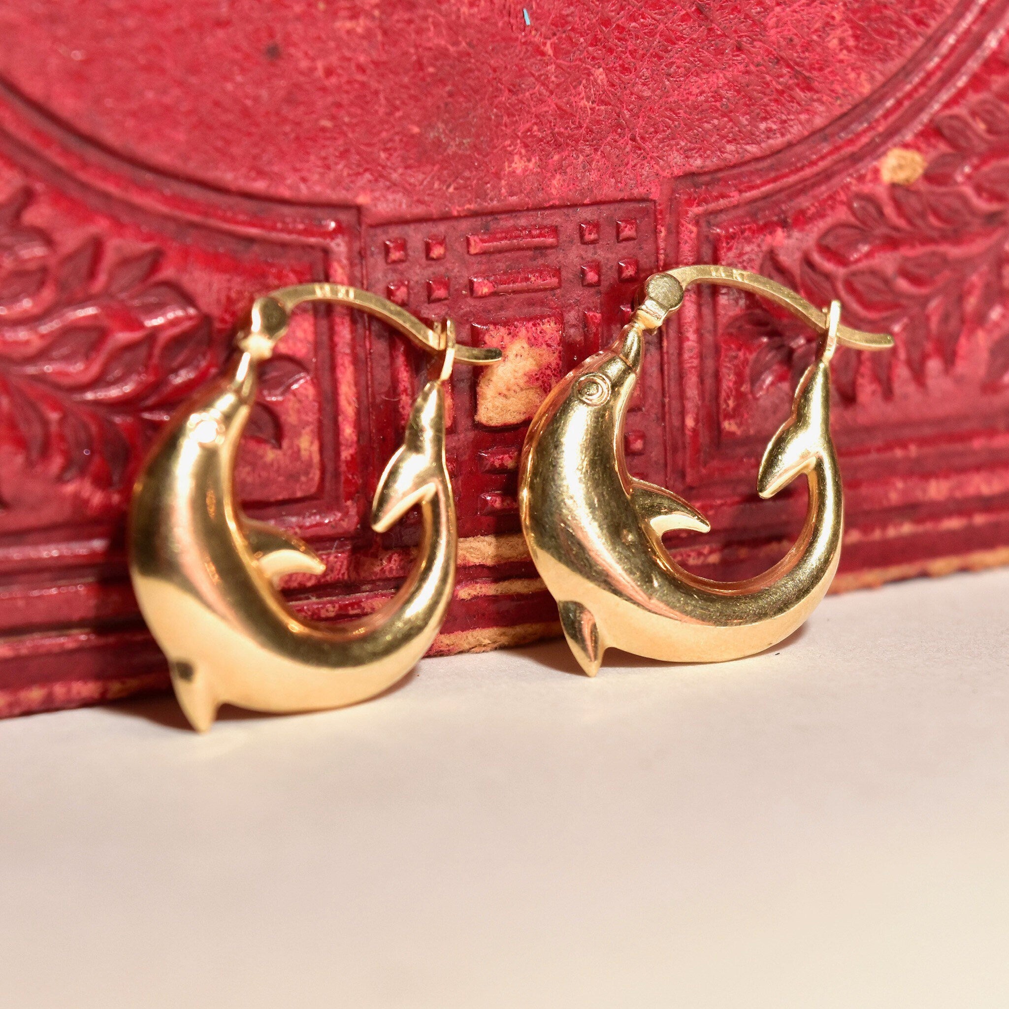 14K yellow gold small dolphin hoop earrings, cute mini oval hoops with leaping dolphin design, 21mm in size, sitting on an ornate red jewelry box.