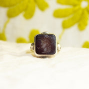 Sterling silver ring with an intaglio black agate carved cameo depicting a Roman warrior profile, displayed on a white textured surface with yellow flower petals in the background.