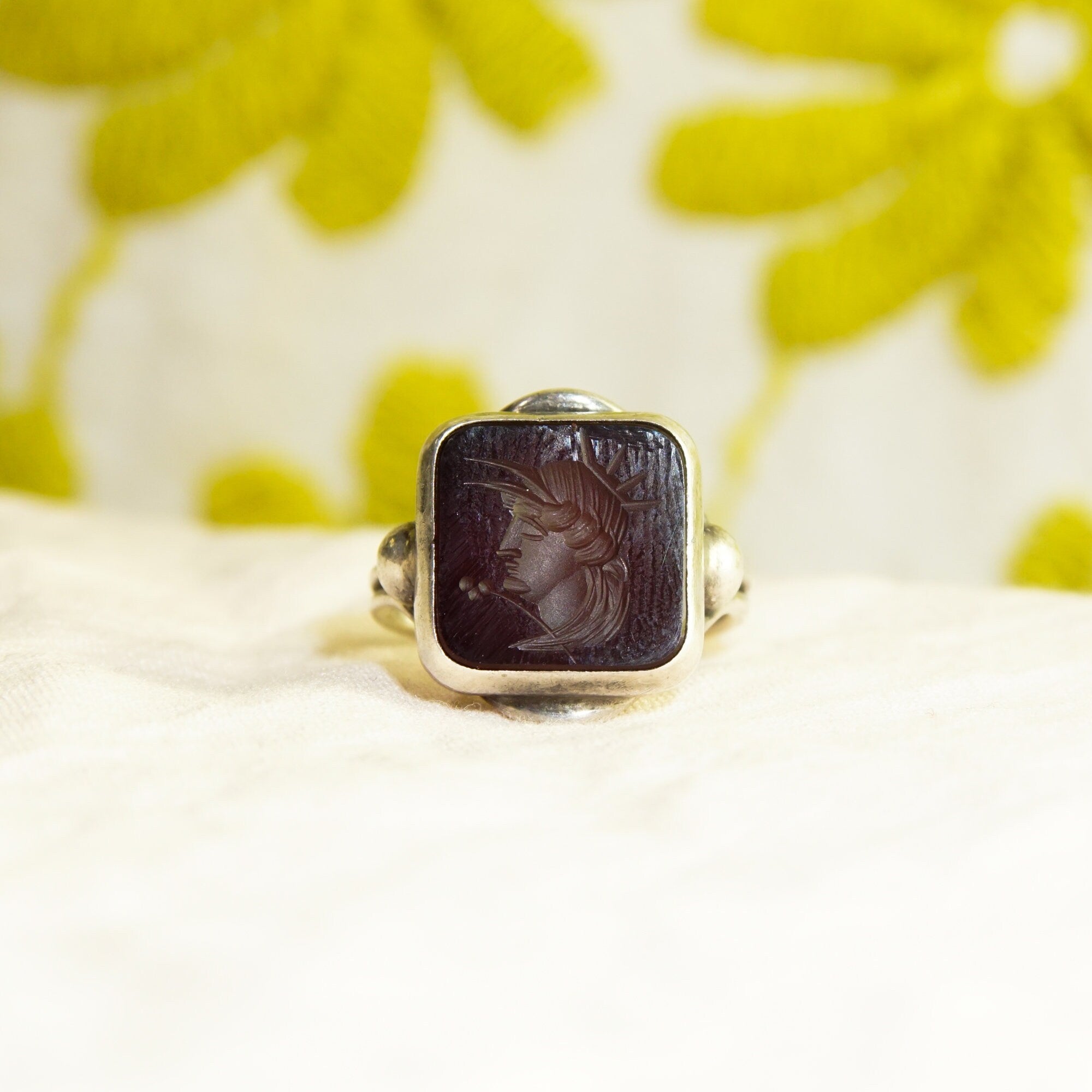 Sterling silver ring with an intaglio black agate carved cameo depicting a Roman warrior profile, displayed on a white textured surface with yellow flower petals in the background.