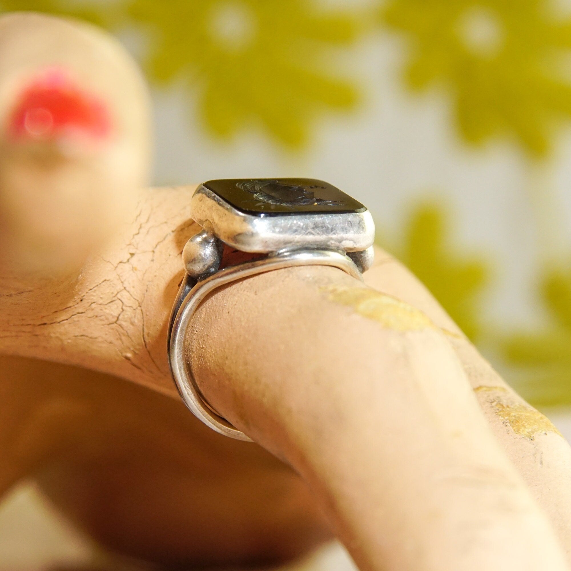 Sterling silver men's pinky ring featuring a carved black agate intaglio seal with a Roman warrior cameo design, shown on a hand with flowers in the background, size 6 US.