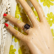 Close-up of a hand wearing a sterling silver men's pinky ring with a carved black agate cameo depicting a Roman warrior, resting on a light-colored knit fabric with green leaf accents, size 6 US.