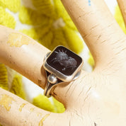 Sterling silver men's pinky ring with carved black agate Roman warrior intaglio cameo seal, size 6 US, showcased on textured beige background with blurred green leaves.