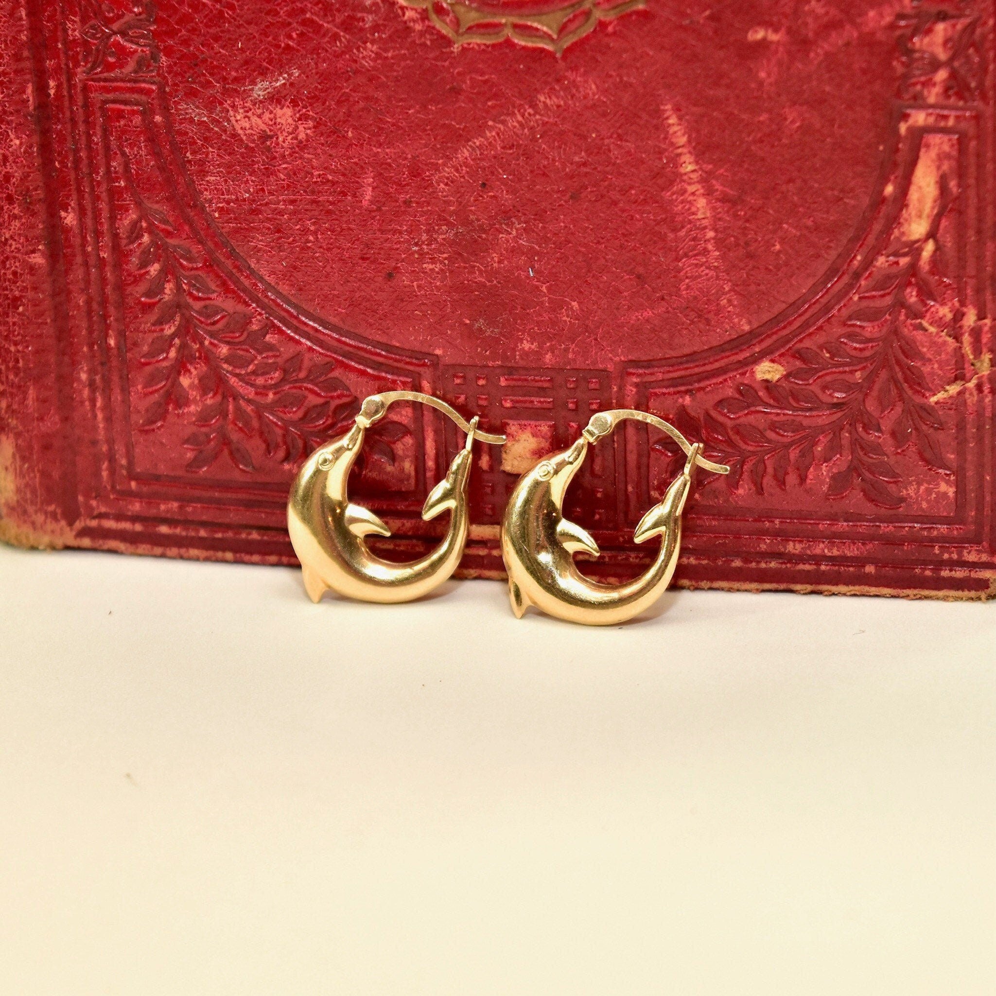 14K yellow gold dolphin hoop earrings lying on antique red book cover, small oval dolphin hoops approximately 21mm in size