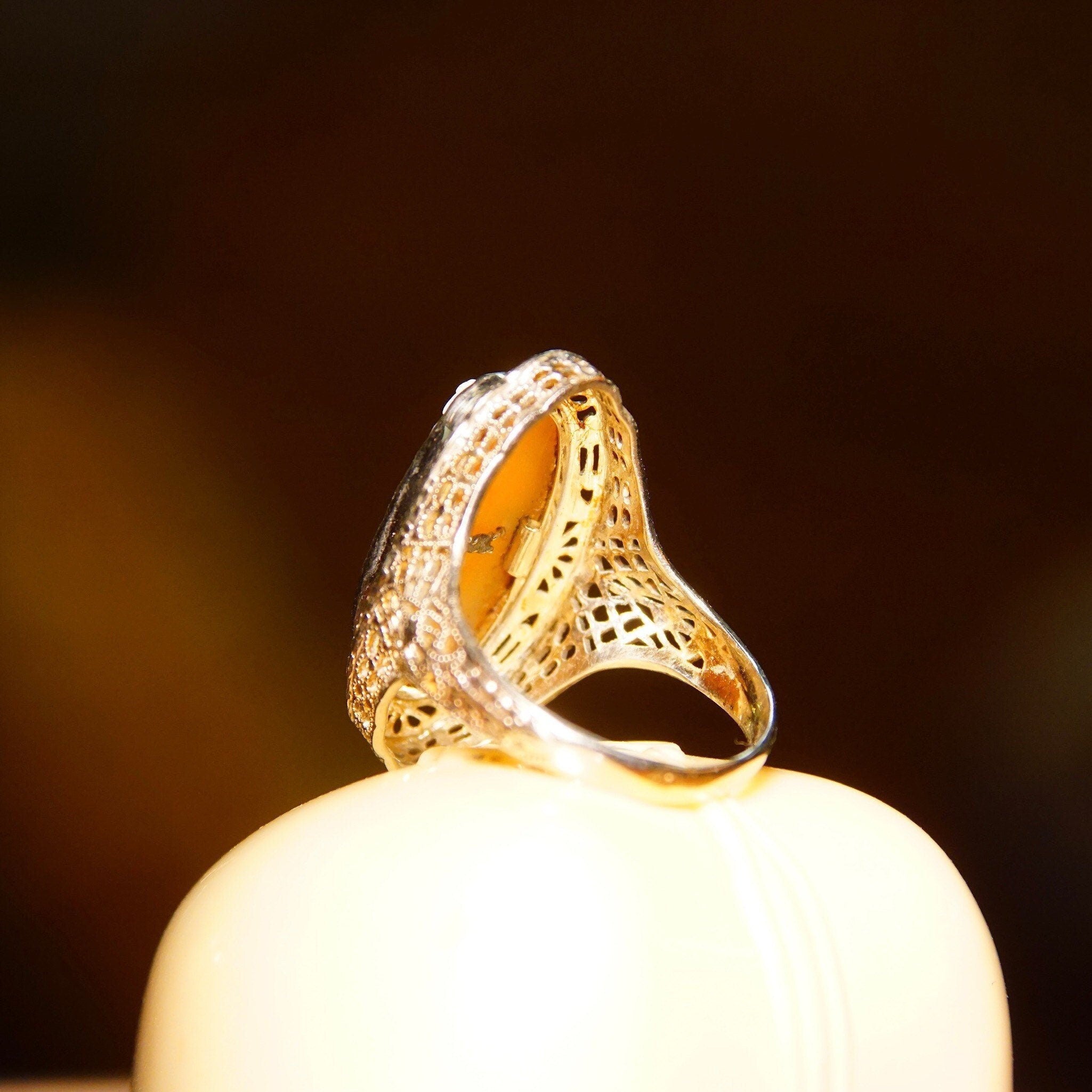 Antique 14K white gold filigree ring with carved shell cameo featuring a woman's bust wearing a diamond necklace, diamond accent on the side of the ring, intricate metalwork detail, size 6 1/4 US by ESEMCO, photographed on a warm brown background.