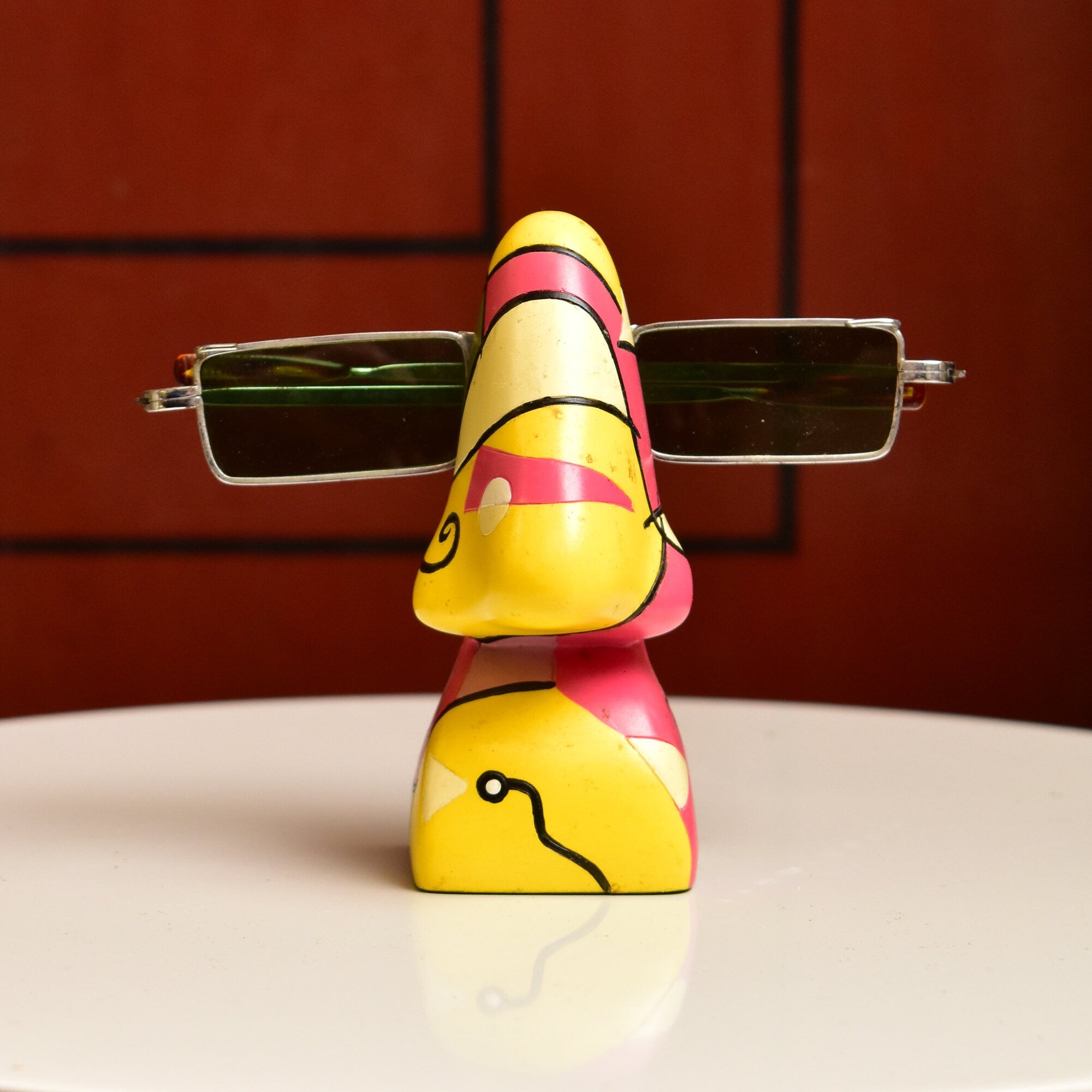 Colorful abstract resin eyeglass holder shaped like a head with Picasso-inspired facial features, including a prominent yellow nose, pink and yellow striped hat, and green spectacles. The vintage home decor piece measures 3 3/4 inches tall and can hold a pair of glasses.