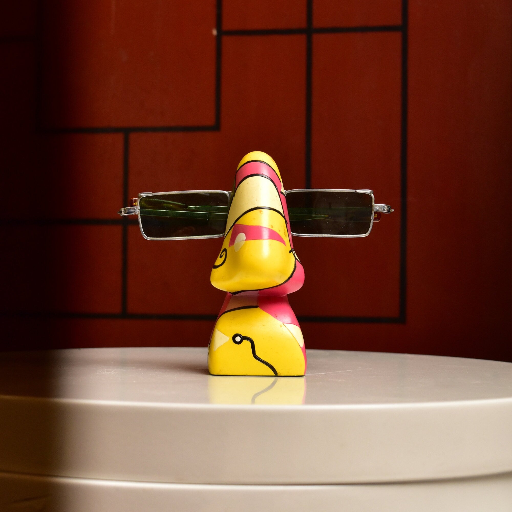 Colorful abstract resin eyeglass stand shaped like a face with exaggerated nose holding glasses, against red brick background, 3 3/4 inches tall, vintage home decor accessory.
