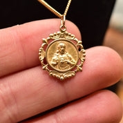 18K gold Sacred Heart of Jesus medallion pendant necklace with embossed religious design, shown held in hand to display intricate detail, on thin gold chain against black background.