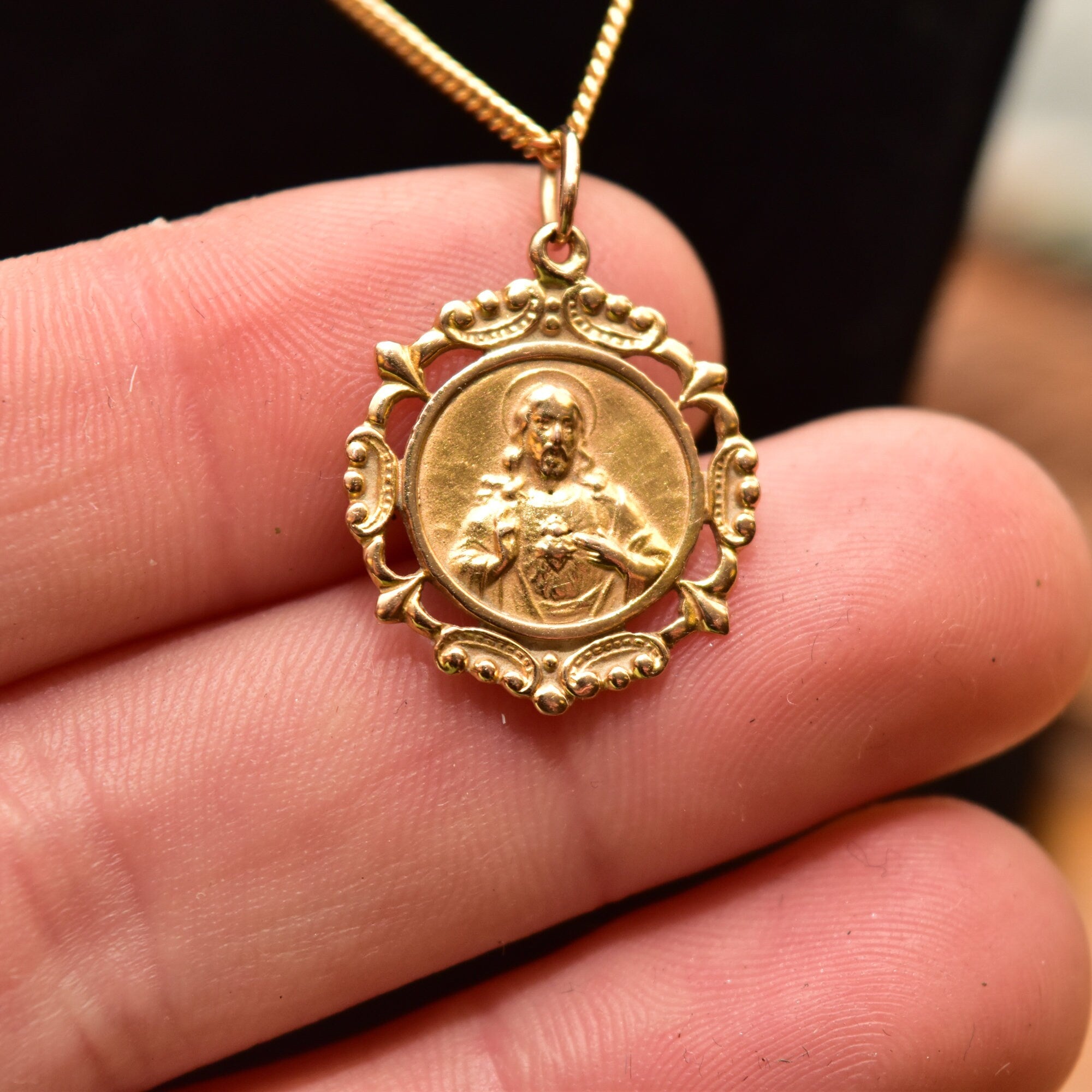 18K gold Sacred Heart of Jesus medallion pendant necklace with embossed religious design, shown held in hand to display intricate detail, on thin gold chain against black background.