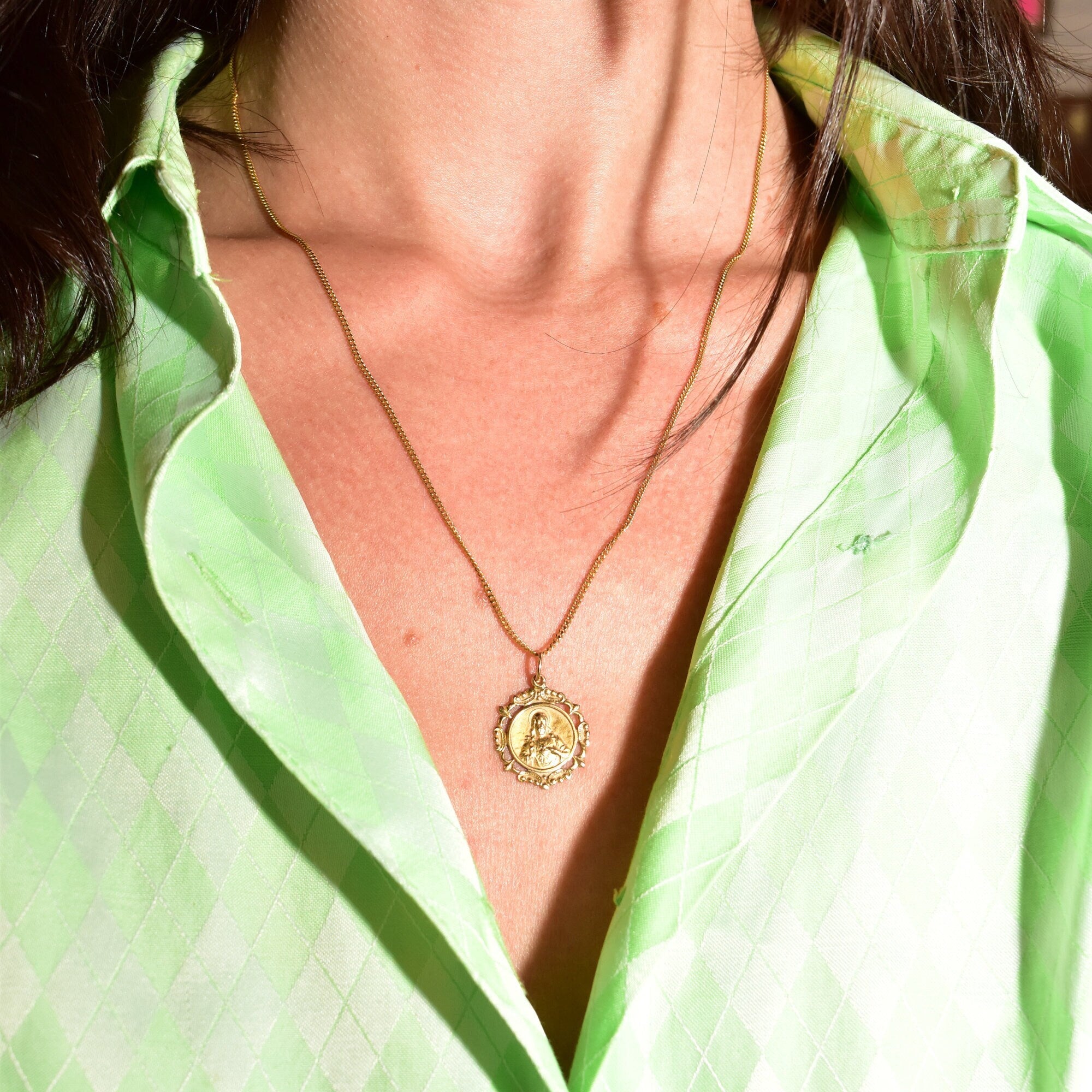 18K yellow gold Sacred Heart of Jesus pendant necklace with embossed medallion on a thin curb chain, worn by a person in a green striped shirt.