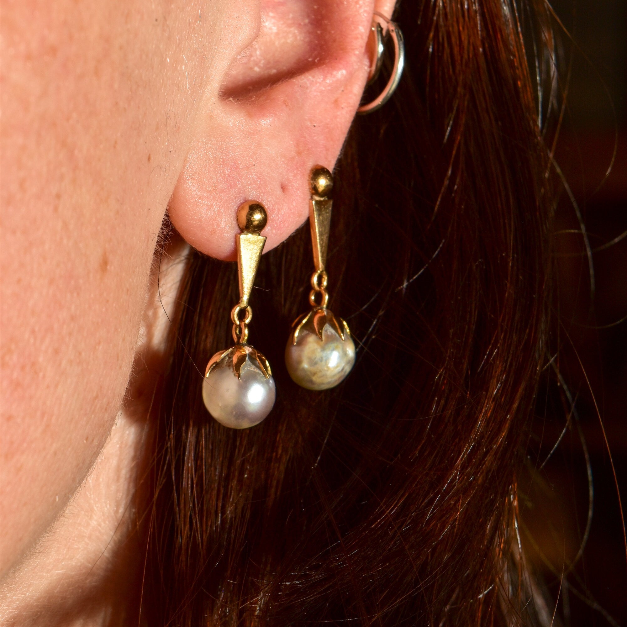 18K yellow gold baroque freshwater pearl dangle earrings shown on a woman's ear, with long brown hair visible