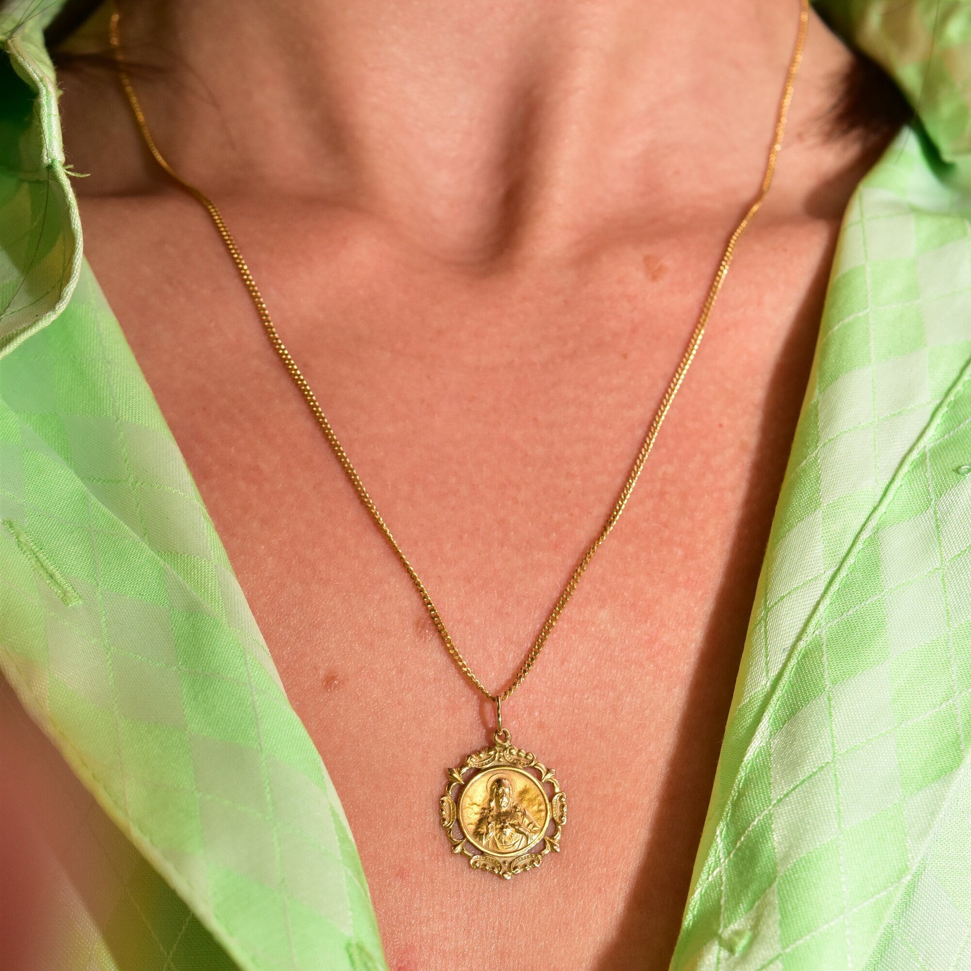 18K yellow gold Sacred Heart Jesus pendant necklace with embossed medallion on a thin curb chain, worn by a person in a green shirt.